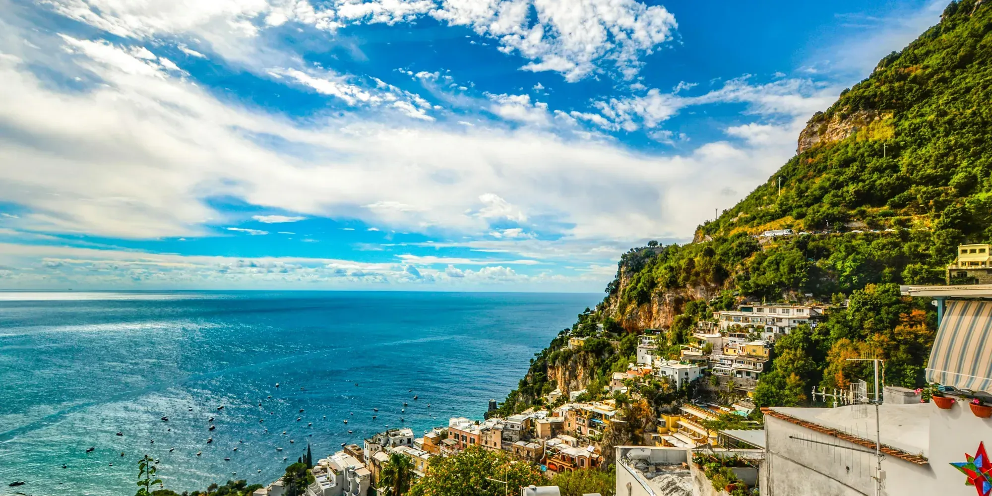 Cliffside buildings overlooking turquoise sea under a blue sky, with lush green hills on a sunny day.