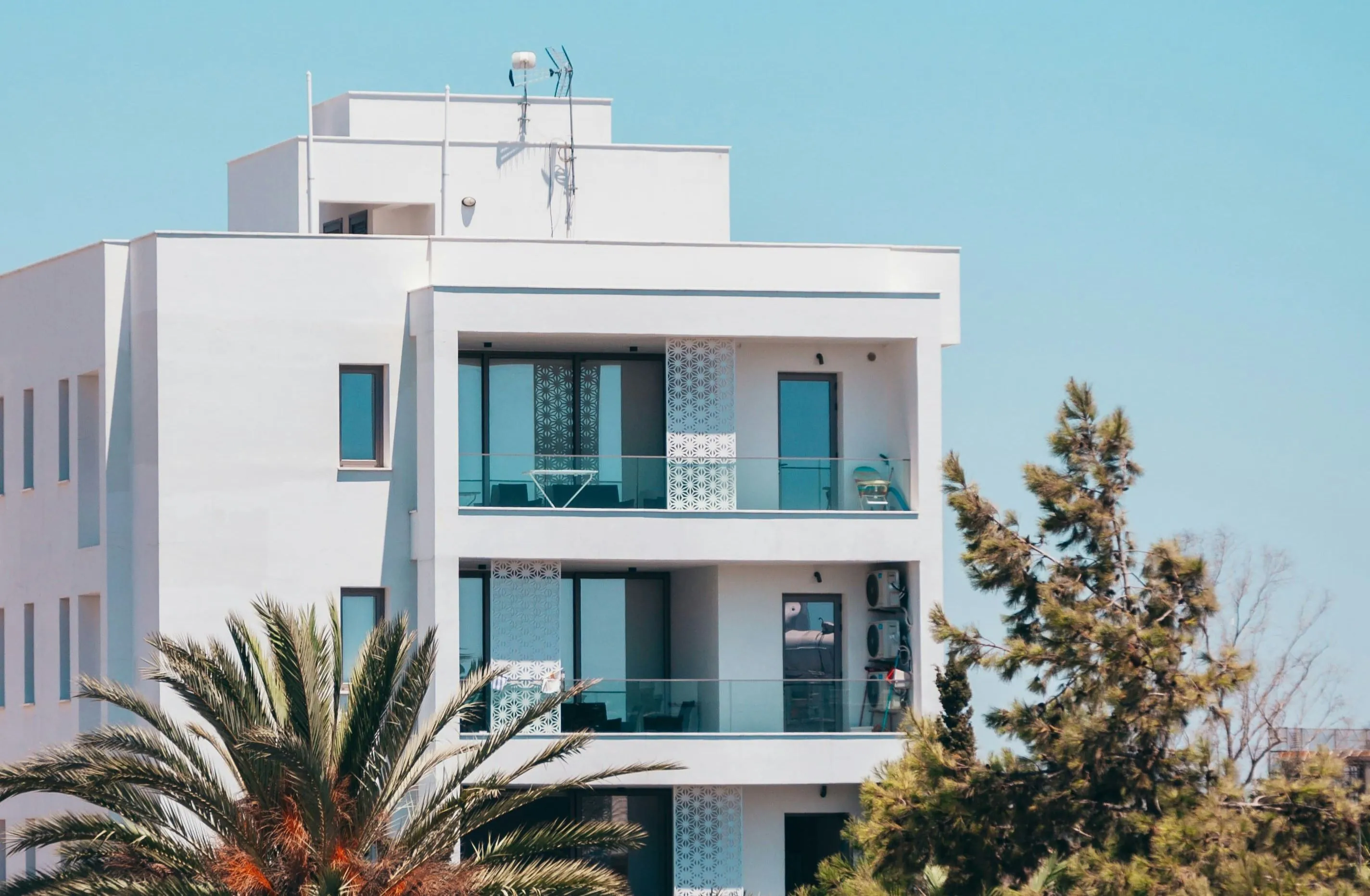 Larnaca Cyprus Real Estate A white, modern apartment building with glass balconies overlooking palm trees and a blue sky.