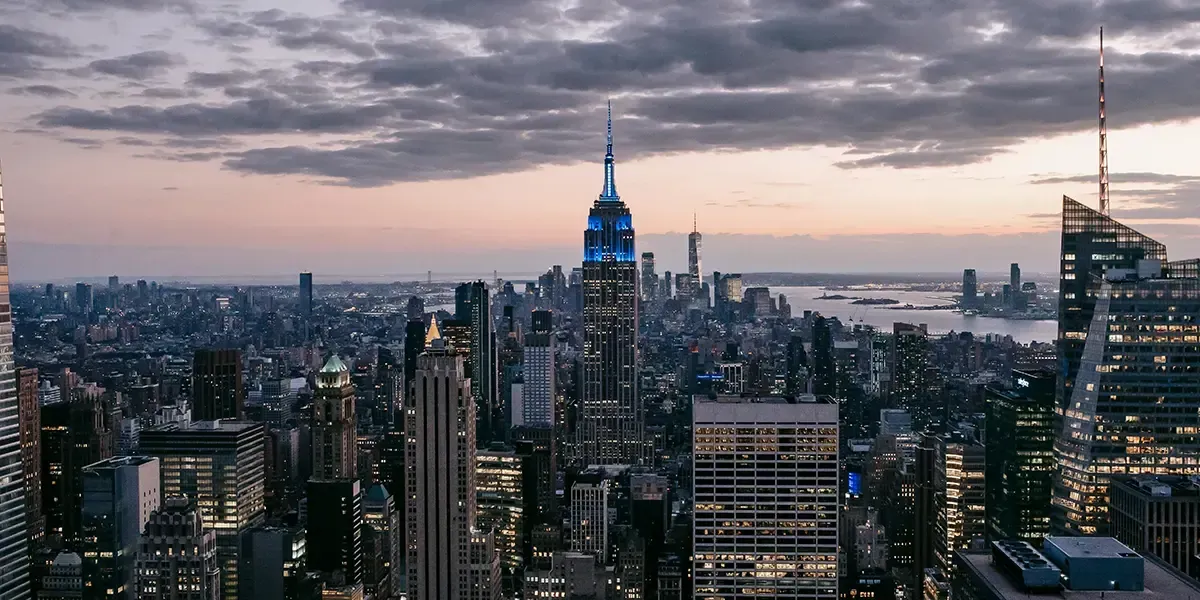 Empire State Building illuminated in blue at dusk, overlooking Manhattan skyline and Hudson River under a cloudy sky